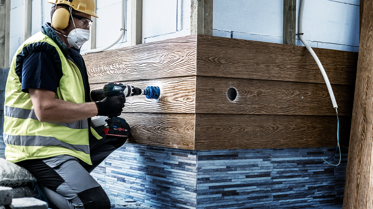 A person wearing safety equipment drills a hole into wooden paneling.