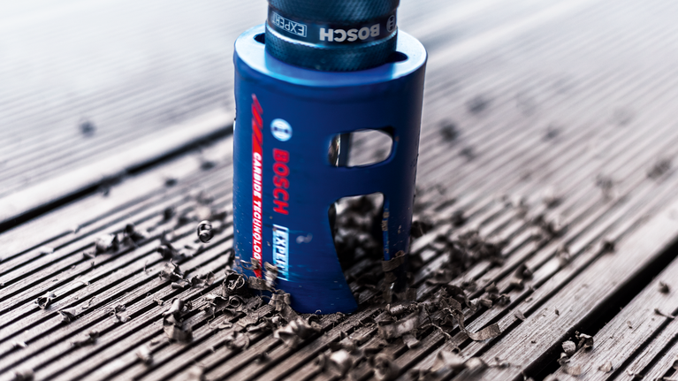 EXPERT Construction Material Hole Saw Set 20/25/32/38/51/64 mm 10-pc Hole saw drilling through a wooden plank, scattering wood shavings.