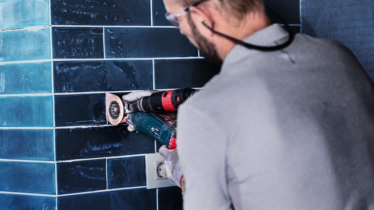 EXPERT 3 max AVZ 70 RT4 Multitool Grouting Plate 70 mm A person wearing safety equipment grinds dark wall tiles with a power tool.