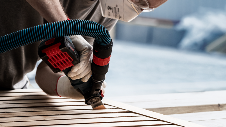 Person wearing safety equipment sands wooden slats with a power tool attached to a vacuum hose.