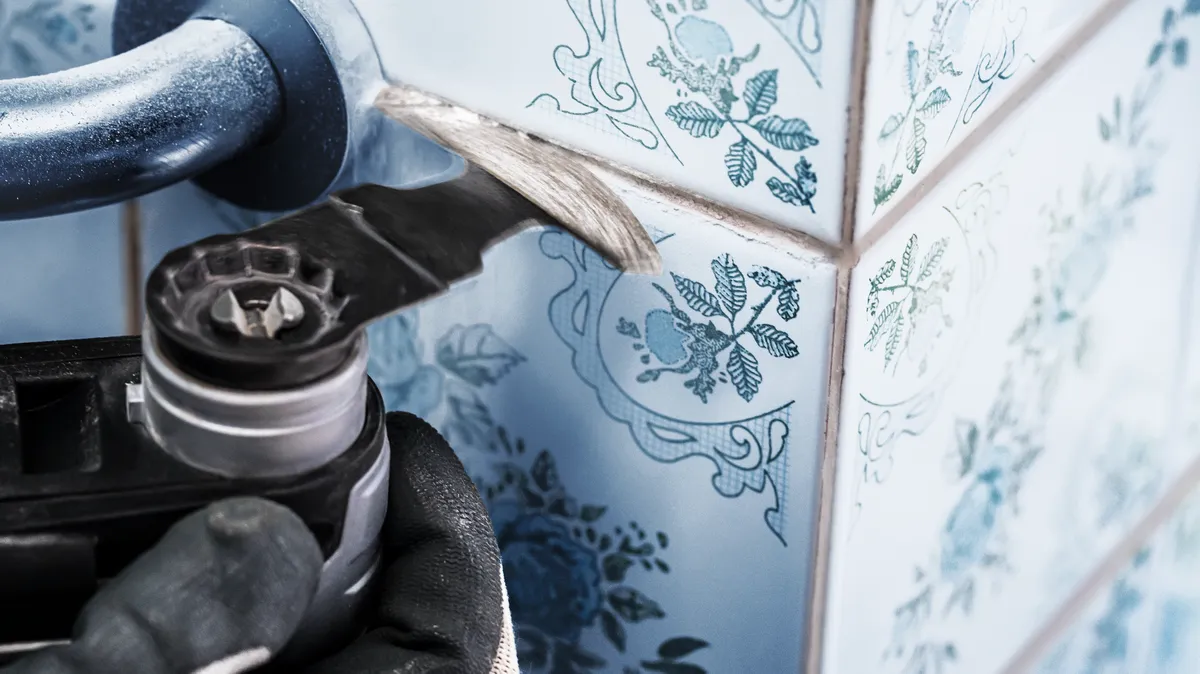Person wearing safety equipment uses a multi-tool to remove grout between floral-patterned tiles.