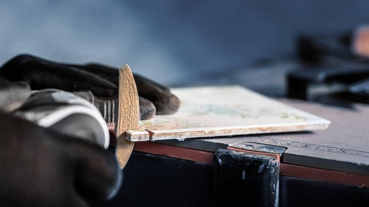 Person wearing safety equipment grinds the edge of a ceramic tile with a power tool.