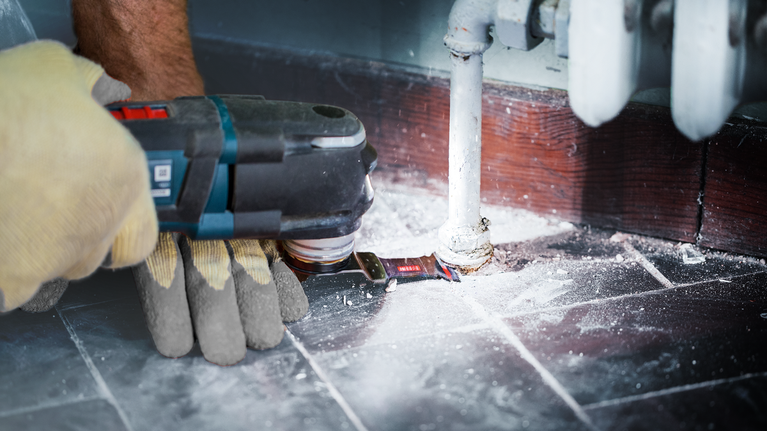 EXPERT MetalMax AIZ 45 AIT Multitool Blade 45 mm A person wearing safety equipment uses a power tool to cut flooring around a pipe.