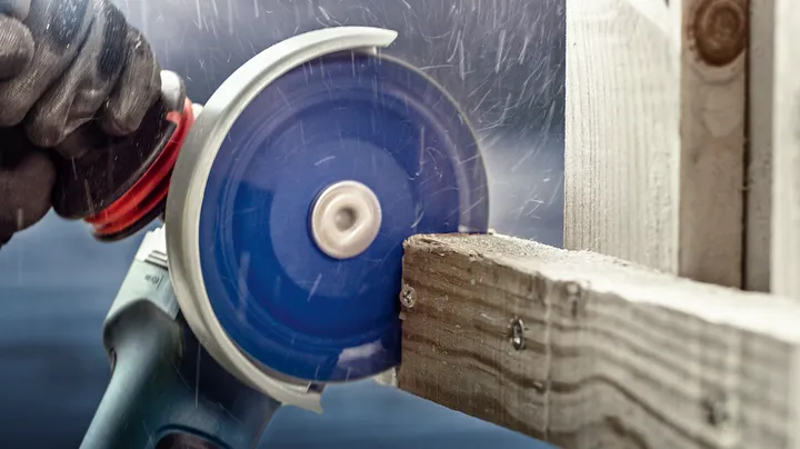 Gloved worker wearing safety equipment cuts a wooden beam with a circular saw.