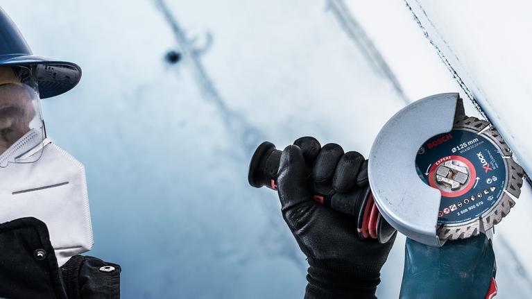 EXPERT MultiMaterial Diamond Cutting Disc 125 x 22.23 x 2.2 x 12 mm A person wearing safety equipment uses an angle grinder to cut a metal surface.