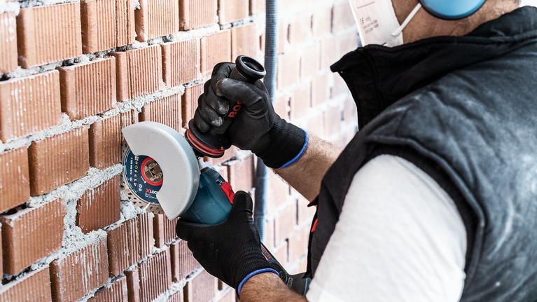 EXPERT MultiMaterial Diamond Cutting Disc 125 x 22.23 x 2.2 x 12 mm Person wearing safety equipment cuts brick wall with an angle grinder.