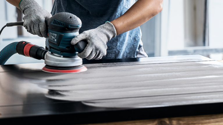EXPERT Multihole Sanding Pad Universal, 150 mm, Medium Person wearing safety equipment sands a wooden surface with a power tool.