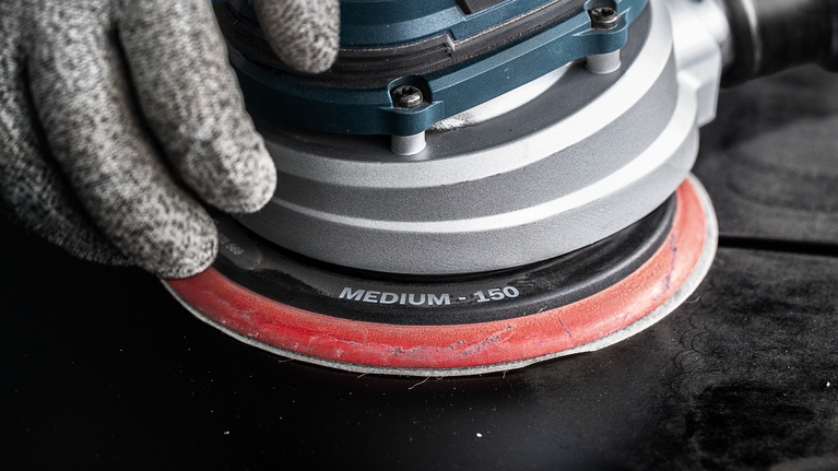 EXPERT Multihole Sanding Pad Universal, 150 mm, Medium Person wearing safety equipment operates a medium-grit orbital sander on a dark surface.