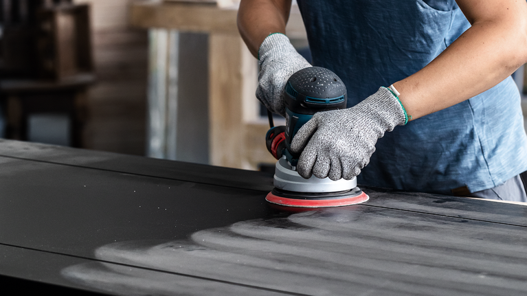EXPERT Multihole Sanding Pad Universal, 150 mm, Medium Person wearing safety equipment sands a wooden surface with a power sander.