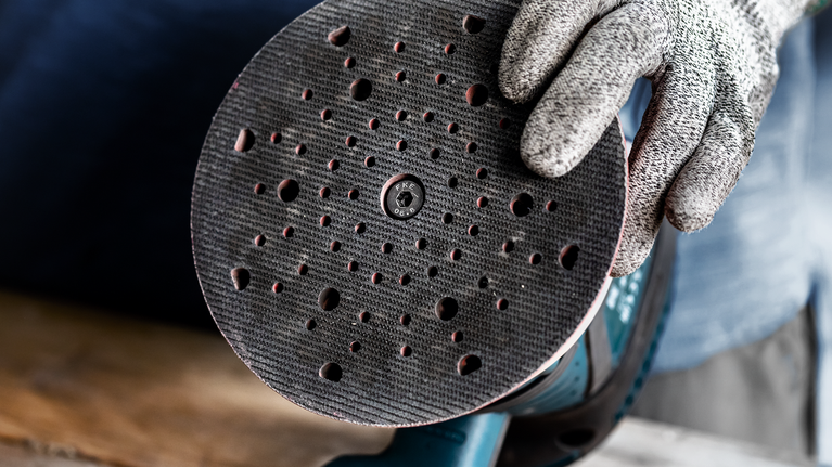 EXPERT Multihole Sanding Pad Universal, 150 mm, Medium Person wearing safety equipment holds a circular sander with a perforated sanding pad.