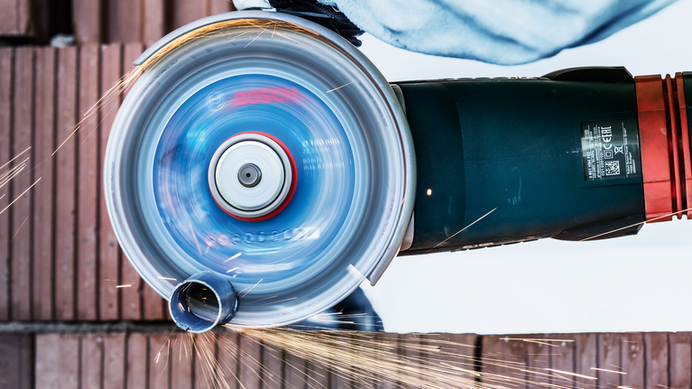 EXPERT Diamond Metal Wheel Cutting Disc 180 x 22.23 mm A person wearing safety equipment cuts a metal pipe with a spinning angle grinder.