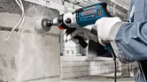 A person wearing safety equipment drills a circular hole into a concrete wall.