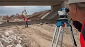 A person wearing safety equipment uses a leveling tool to survey a construction site.