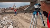 Person wearing safety equipment uses a surveying level tripod on a construction site.