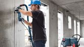 A person wearing safety equipment drills into concrete with a dust extractor connected.