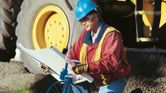 A person wearing safety equipment checks blueprints beside a measuring wheel at a construction site.