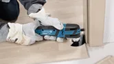 A person wearing safety equipment cuts laminate flooring with a cordless multi-cutter.
