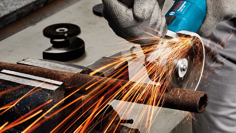 A person wearing safety equipment grinds a rusted metal pipe with an angle grinder.