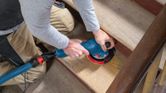 A person sands wooden stairs with a random orbit sander connected to a vacuum hose.