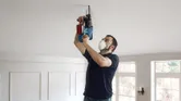 A person wearing safety equipment drills into a ceiling with a power tool.