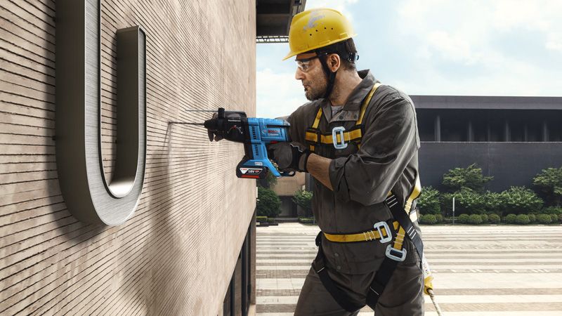 A person wearing safety equipment drills into a building wall while suspended.