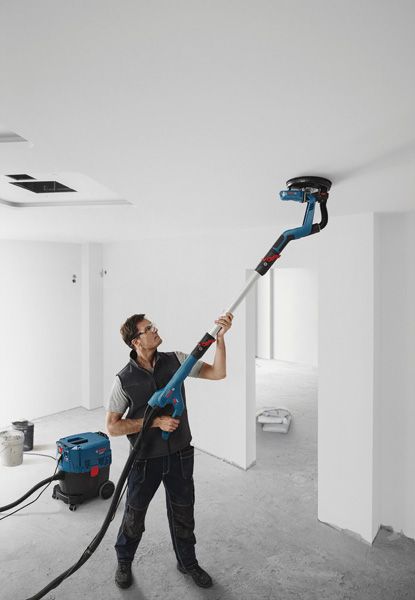 A person wearing safety equipment sands a ceiling with a drywall sander in an unfinished room.