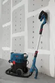 Drywall sander connected to a dust extractor standing against an unfinished wall.