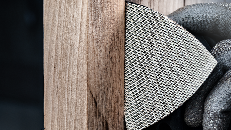 Person wearing safety equipment sands a wooden surface with a mesh sanding pad.