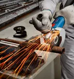 A person wearing safety equipment grinds a metal pipe with an angle grinder.