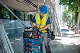 A person wearing safety equipment organizes a tool bag on stacked tool cases at a construction site.