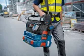 Person wearing safety equipment carries stacked toolboxes and a digital angle measurer at a construction site.
