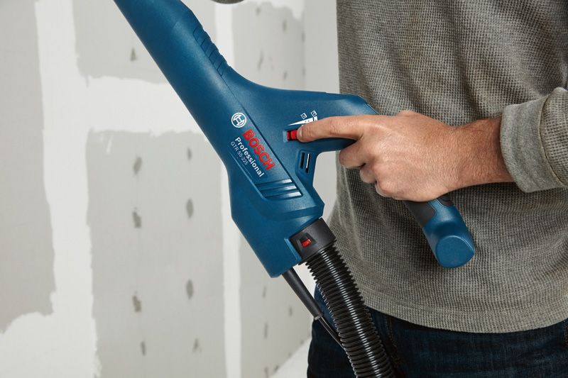 A person adjusts a blue power tool with a hose near an unfinished drywall.