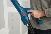 A person adjusts a blue power tool with a hose near an unfinished drywall.