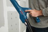 A person adjusts a blue power tool with a hose near an unfinished drywall.