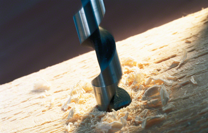 Drill bit boring a hole into a wooden surface with wood shavings around.