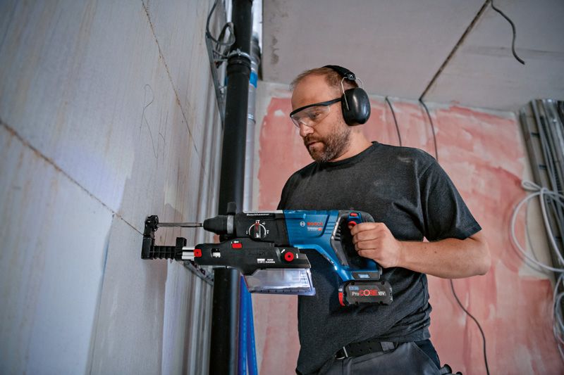 A person wearing safety equipment drills into a wall using a tool with an integrated dust extractor.