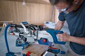 A person wearing safety equipment cuts wood with a cordless mitre saw on a workbench.
