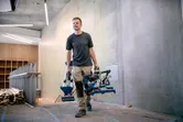 A person carries a cordless mitre saw and stand in a building under construction.