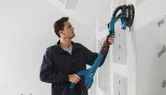 A person sands drywall with a power drywall sander on an unfinished wall.