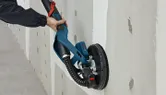 A person sands drywall with a power sander attached to a dust extraction hose.