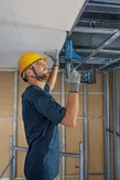 A person wearing safety equipment fastens metal ceiling framework with a power tool.