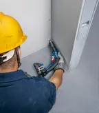 A person wearing safety equipment drives screws into drywall with a power tool.