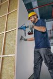 A person wearing safety equipment secures drywall to a frame using an automatic screw gun.