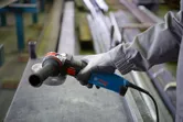 A person wearing safety equipment holds an angle grinder near a metal surface.