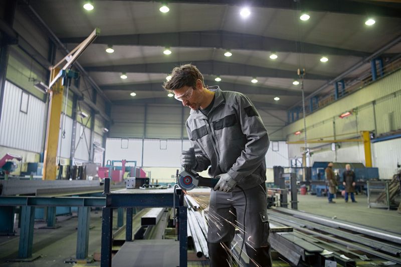 A person wearing safety equipment grinds metal, sparks flying in a large industrial workshop.