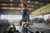 A person wearing safety equipment carries an angle grinder in a large industrial workshop.