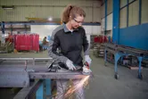 A person wearing safety equipment uses an angle grinder to cut metal, creating sparks.