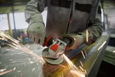 A person wearing safety equipment grinds metal with an angle grinder, producing sparks.