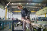 A person wearing safety equipment uses an angle grinder to cut metal in a factory.