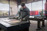 A worker wearing safety equipment grinds metal on a large workbench in a factory.